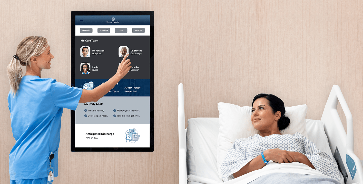 Nurse using a large bedside touchscreen to review care team and daily goals while a patient in a hospital bed looks on, illustrating digital patient engagement technology.