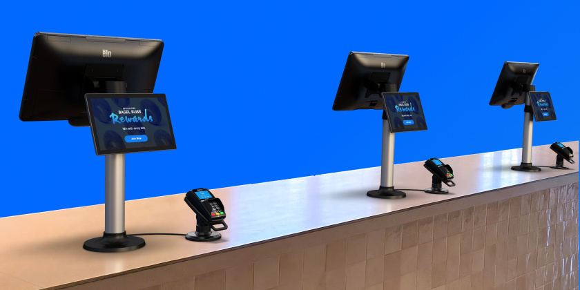 Multiple Elo touchscreen displays mounted on stands along a retail checkout counter, each showing digital promotional content, with payment terminals placed in front to support in-store retail media and POS interactions.